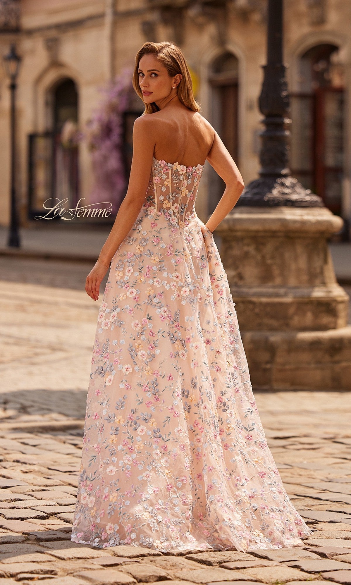 A woman stands on a cobblestone street wearing the La Femme 33409 long prom dress—a light pink floral gown featuring a strapless sweetheart neckline and a sheer, corset-style back.